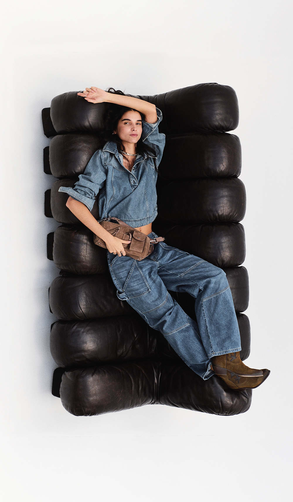 A model reclining on a black sofa wearing jeans and a blue blouse with a bag and Curtis boots by BA&SH.