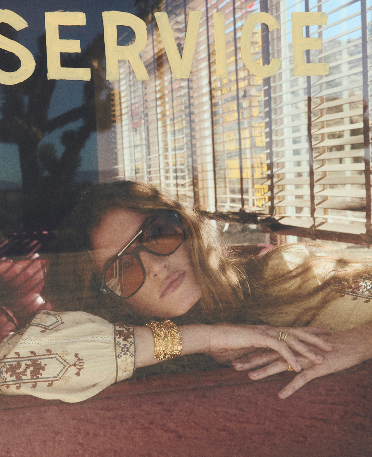 A model wearing a cream embroidered blouse with brown sunglasses. She is sitting on a diner bench.
